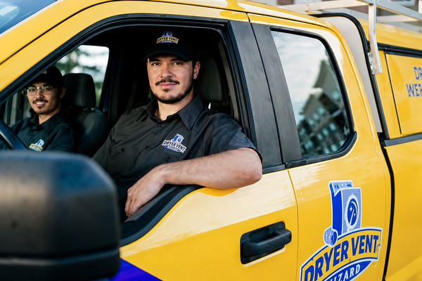 Two men in black uniforms with Dryer Vent Wizard logos sit in a bright yellow service truck.