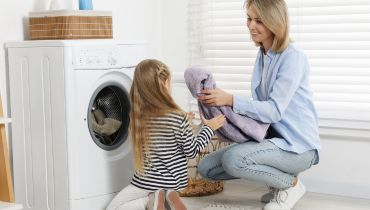 A woman and a young girl are in a laundry room. The woman is handing a purple towel to the girl, who is kneeling near a washing machine.
